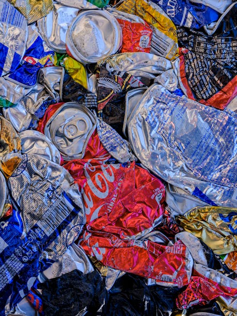 A close-up view of a pile of crushed aluminium beverage cans, including soda cans with various colours such as silver, red, blue, black, and gold. The cans are crumpled and stacked irregularly, with s
